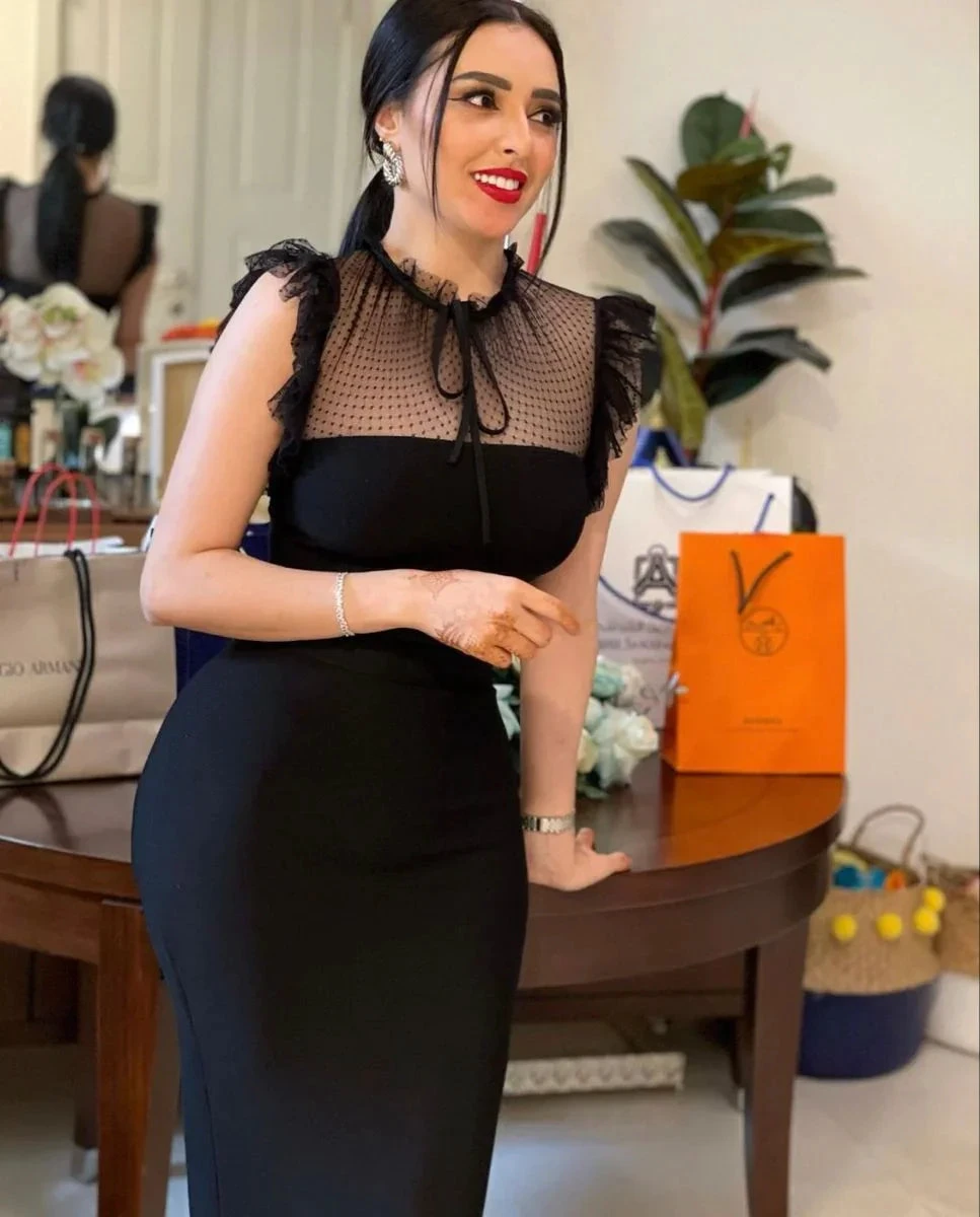 Woman in elegant black dress with sheer polka-dot top, standing indoors near shopping bags, ZAPDRESS fashion