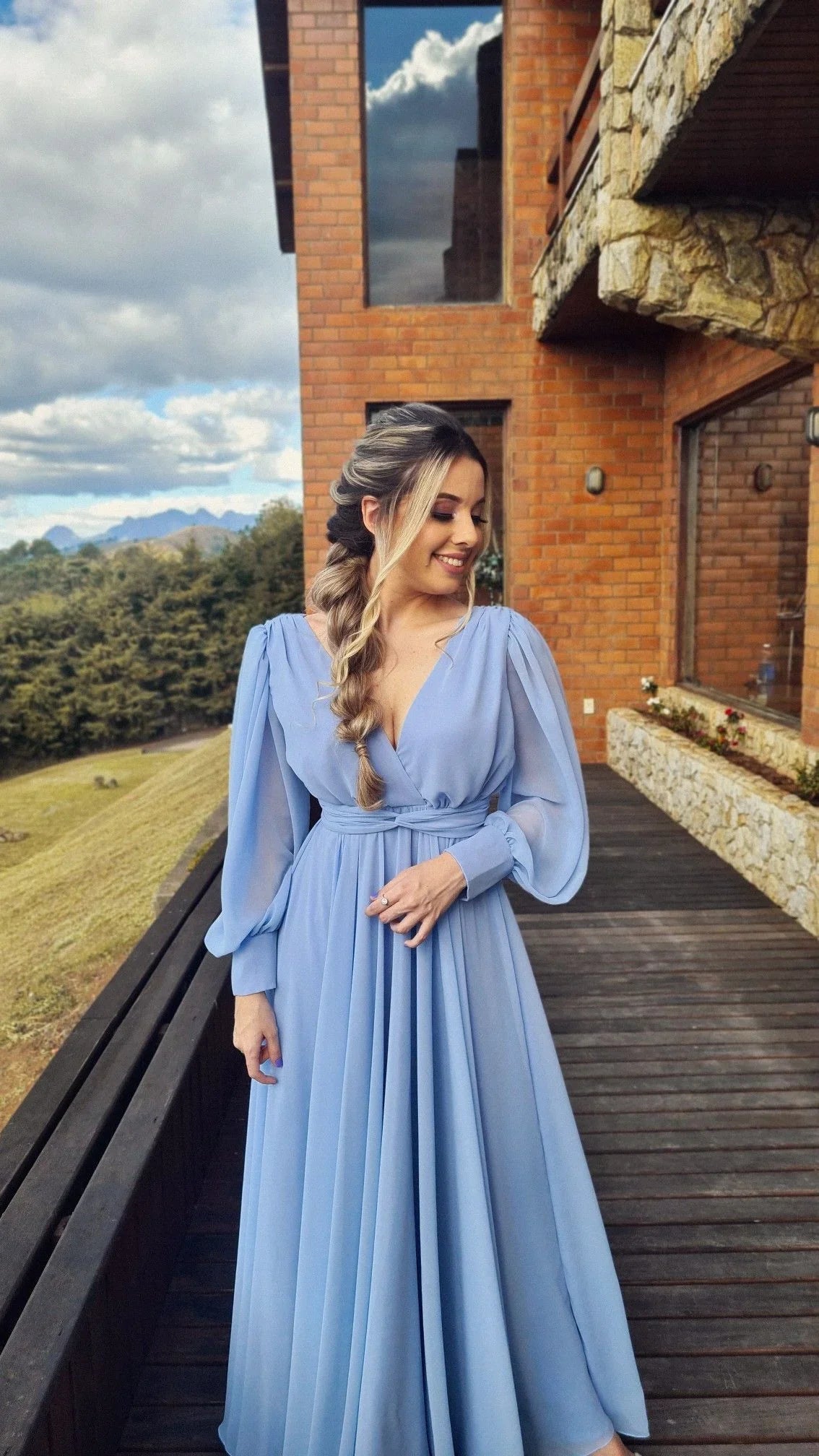 Woman in light blue long dress standing on wooden deck by brick house, ZAPDRESS fashion