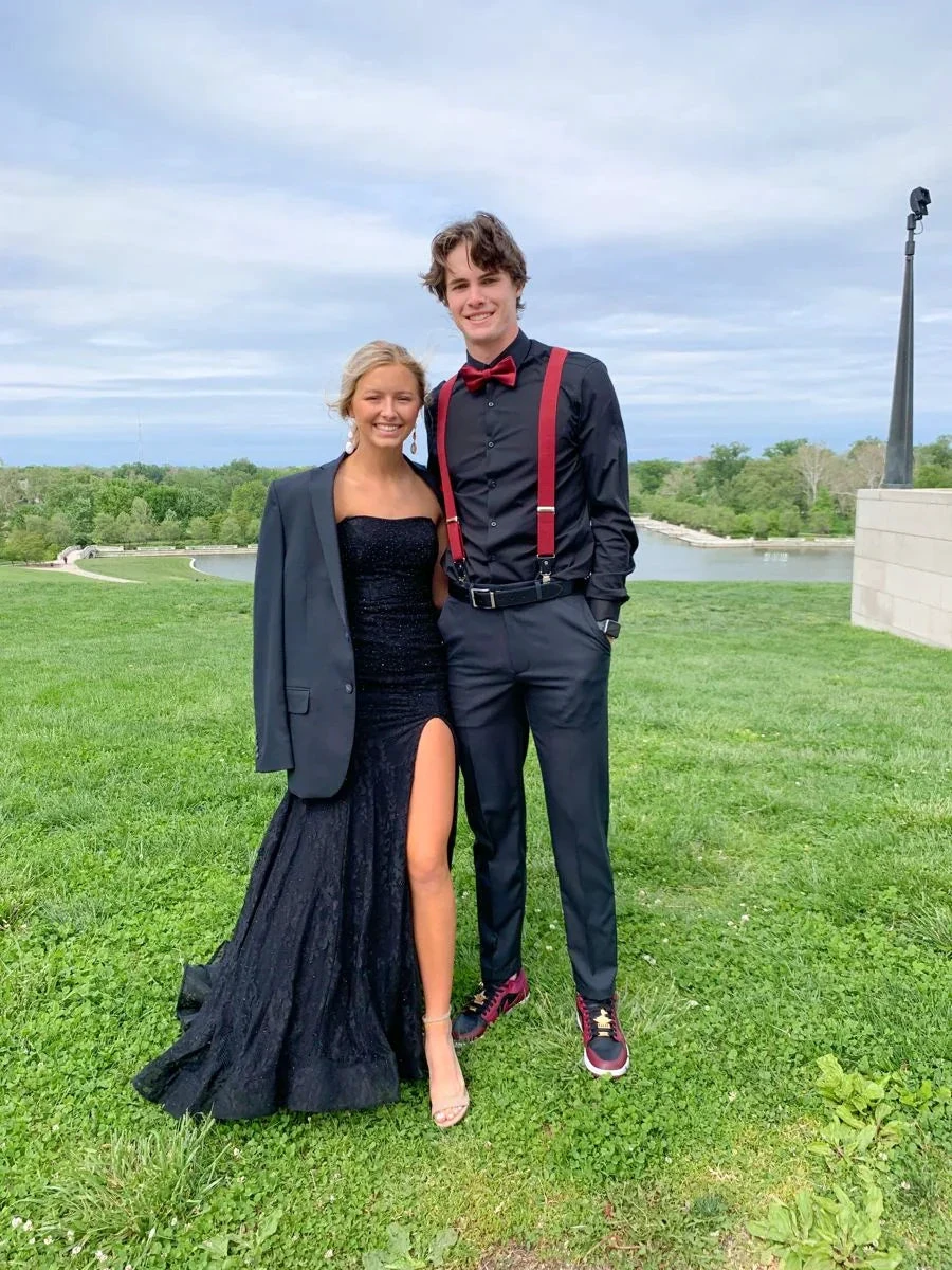 Young couple in prom outfits, woman in black evening dress with jacket, man in black shirt with red suspenders and bow tie, standing on grass with scenic background, ZAPDRESS style
