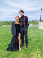 Young couple in prom outfits, woman in black evening dress with jacket, man in black shirt with red suspenders and bow tie, standing on grass with scenic background, ZAPDRESS style