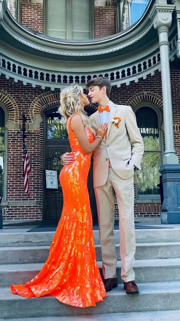 Couple in formal attire, woman in a bright orange mermaid prom dress by ZAPDRESS, standing on historic building steps