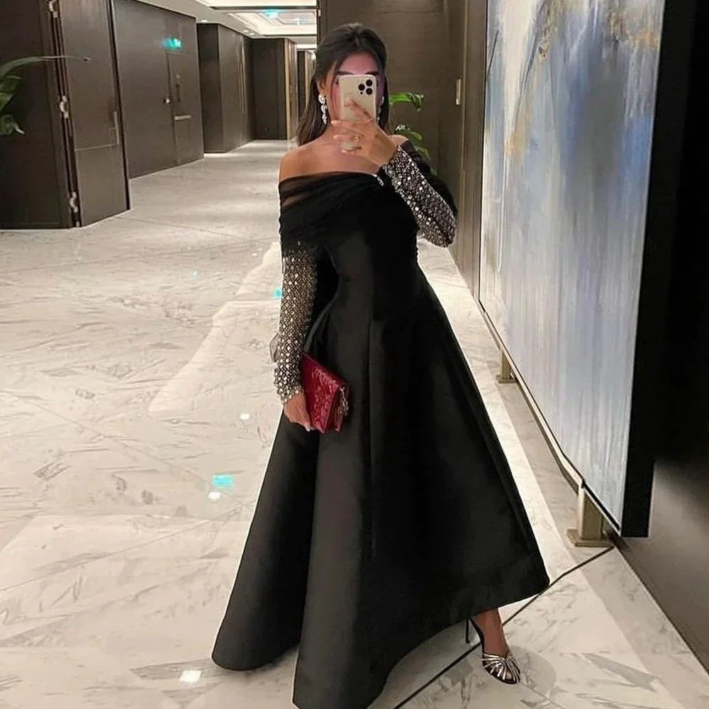 Elegant woman in a black off-shoulder evening dress with sheer sequin sleeves, holding a red clutch, in a modern hallway. ZAPDRESS formal fashion.