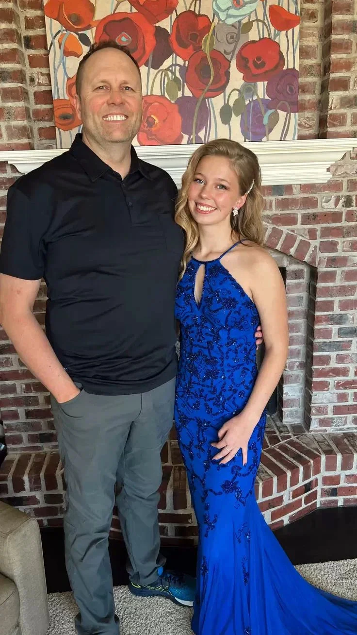 Smiling woman in elegant royal blue gown beside man in black shirt, standing indoors by brick fireplace and floral art