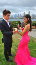 Couple in formal attire exchanging corsages, woman in hot pink prom dress by waterfront city skyline