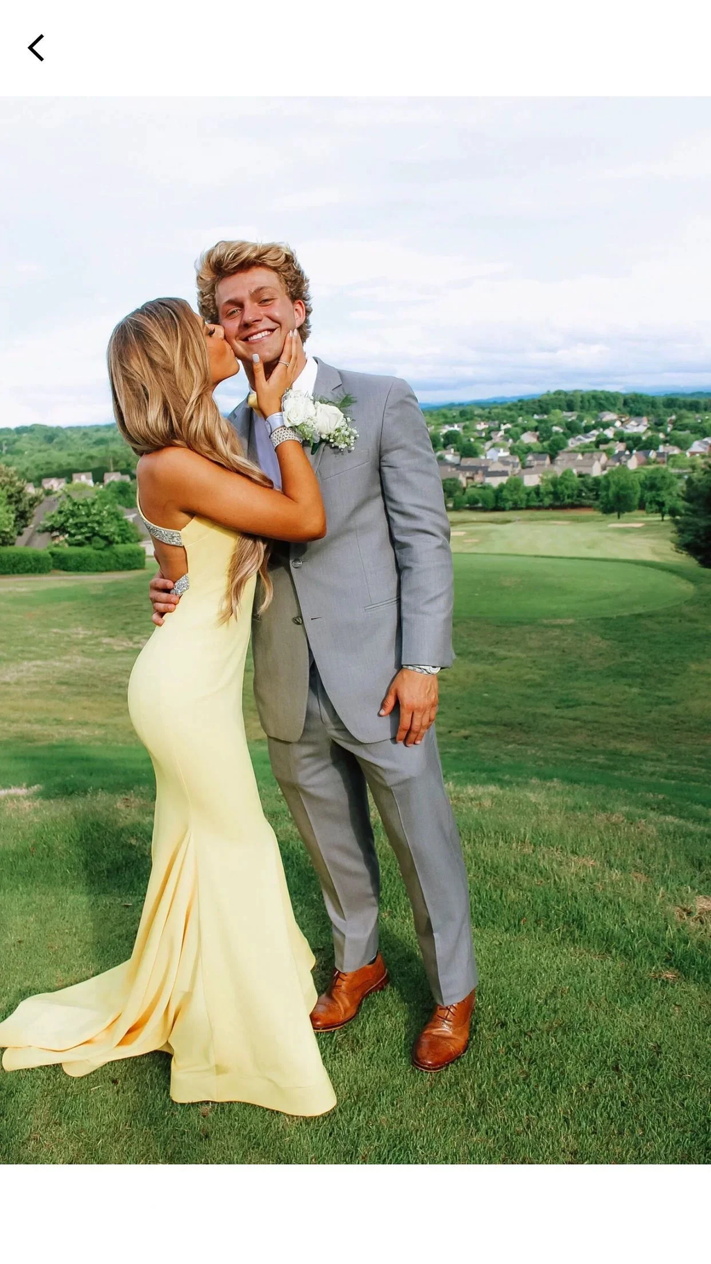 Woman in yellow prom dress kisses man in gray suit on grassy hill, ZAPDRESS formalwear