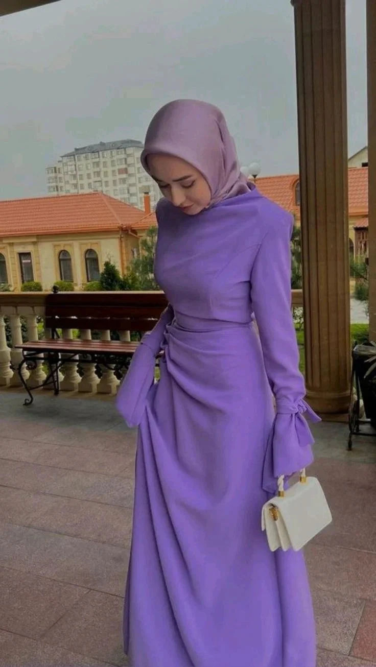 Woman wearing elegant lilac modest dress and matching hijab, holding white handbag outdoors