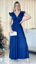 Woman in a royal blue maxi dress holding a white clutch, standing indoors near a sofa, ZAPDRESS fashion.