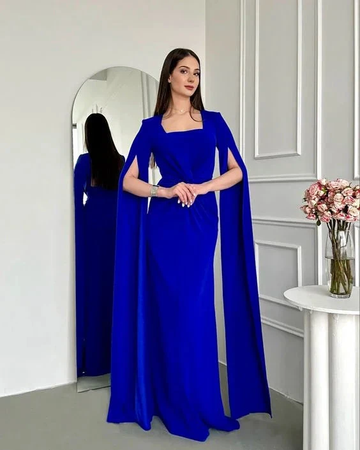 Woman in royal blue evening gown with cape detail, standing indoors near a mirror, ZAPDRESS fashion.