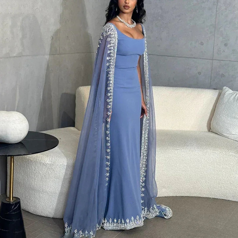 Woman wearing elegant blue embellished evening dress with cape, standing in modern living room