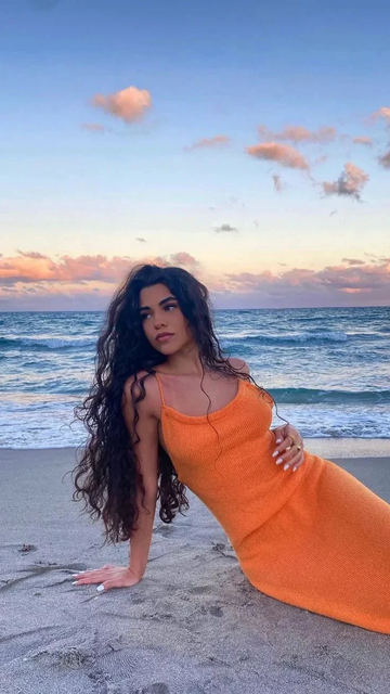 Woman in orange dress sitting on sandy beach at sunset, ocean waves in background, ZAPDRESS style