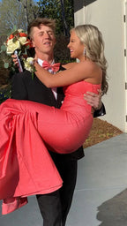 Man in black tuxedo holding smiling woman in sparkling coral dress, outdoor daytime event