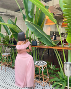 Woman in a pink off-shoulder dress sitting indoors at a tropical-themed café with large green plants, trendy bar stools, and modern decor, ZAPDRESS fashion style.