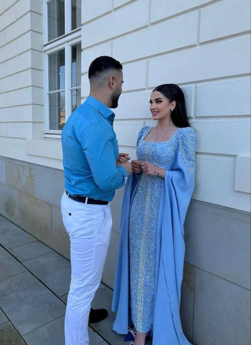Elegant woman in ornate blue gown and man in blue shirt, standing outdoors by ZAPDRESS store wall
