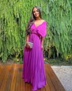 Woman in long magenta dress holding clutch on wooden deck, green fern backdrop, ZAPDRESS fashion