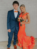 Young couple in formal attire, woman in bright orange sequin gown, posing by white brick wall