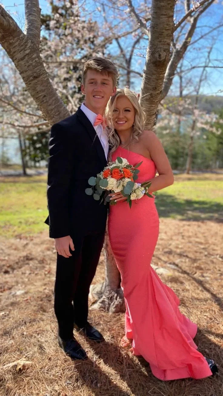 Couple in formal attire, woman in coral pink gown holding bouquet, standing by tree outdoors