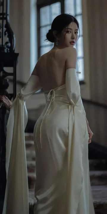 Elegant woman in backless ivory gown with long sleeves, posing on a staircase, ZAPDRESS