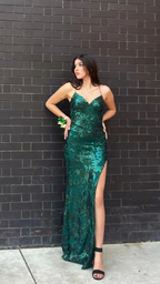 Woman in emerald green sequin evening gown with a side slit, posing by a black brick wall, ZAPDRESS