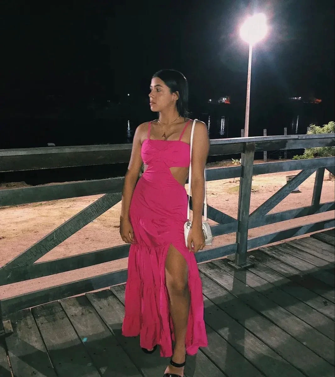 Woman in bright pink sleeveless dress with side cutout, standing on wooden boardwalk at night