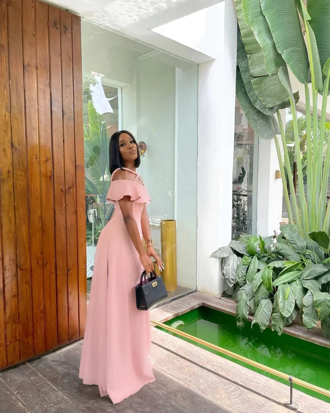 Woman in a pink off-shoulder dress holding a black handbag by indoor plants at ZAPDRESS