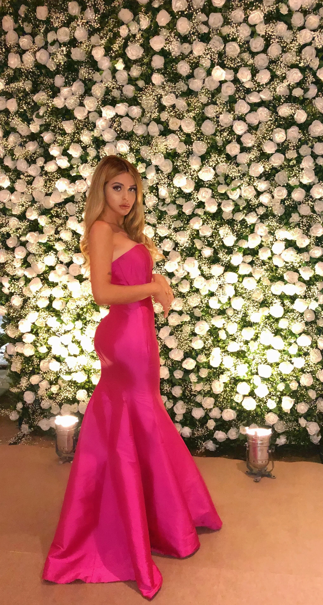 Woman in a bright pink evening gown posing before a white rose flower wall, ZAPDRESS style