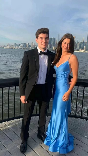 Young couple in formal attire by waterfront, woman in blue satin dress from ZAPDRESS, city skyline view
