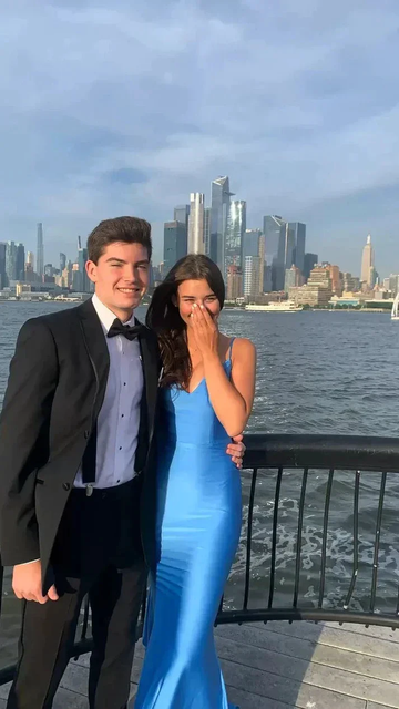 Couple in formal attire by waterfront with city skyline, woman in blue ZAPDRESS gown